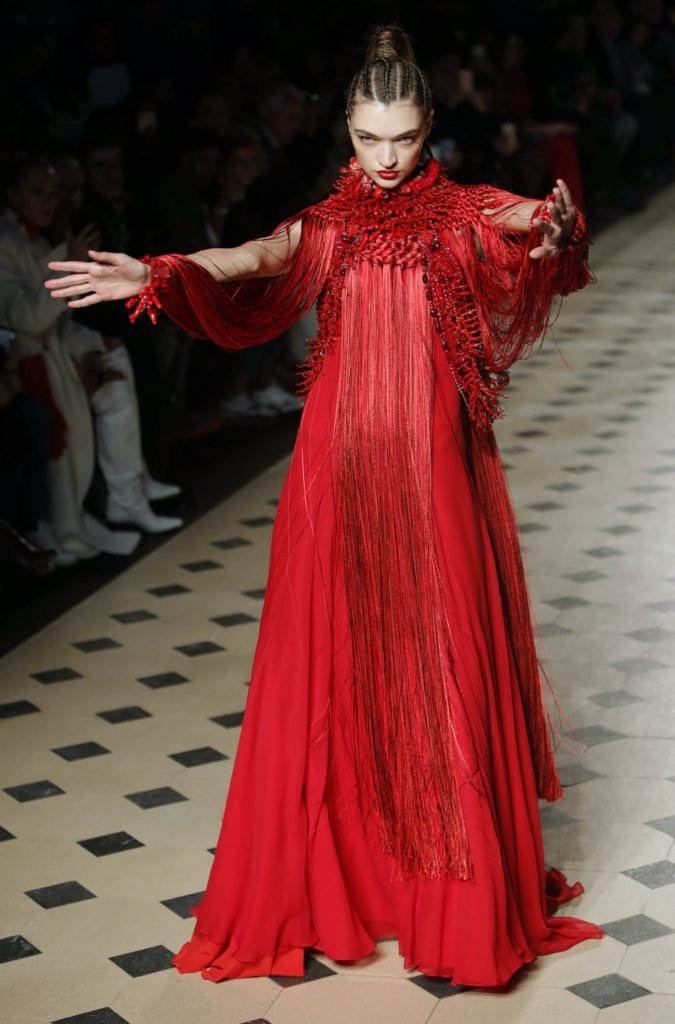 Julien Fournié défilé Haute Couture, un mannequin habillé d'une robe rouge déploie ses bras et offre un regard mystérieux aux spectateurs.